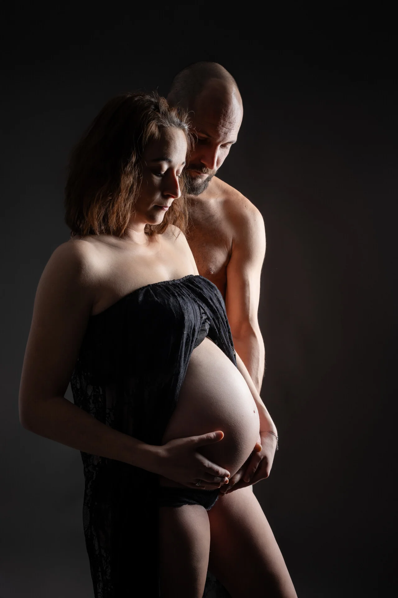 Future maman et papa en couple, en robe noire, lumière artistique en contre-jour – séance grossesse en studio près de Rouen
