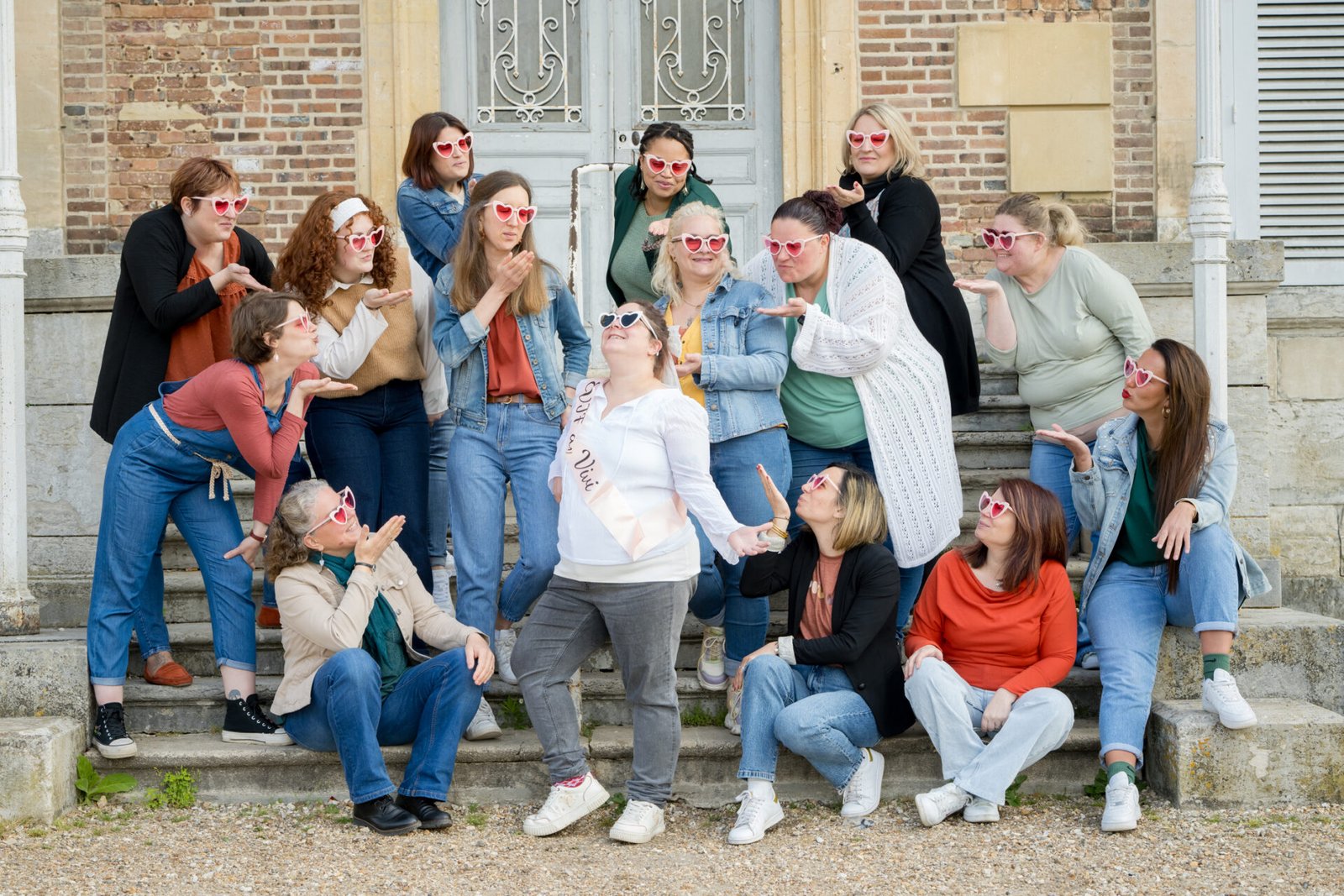 Groupe de femmes en lunettes cœur célébrant un EVJF avec joie, posant sur les marches d’un bâtiment à l’ambiance vintage