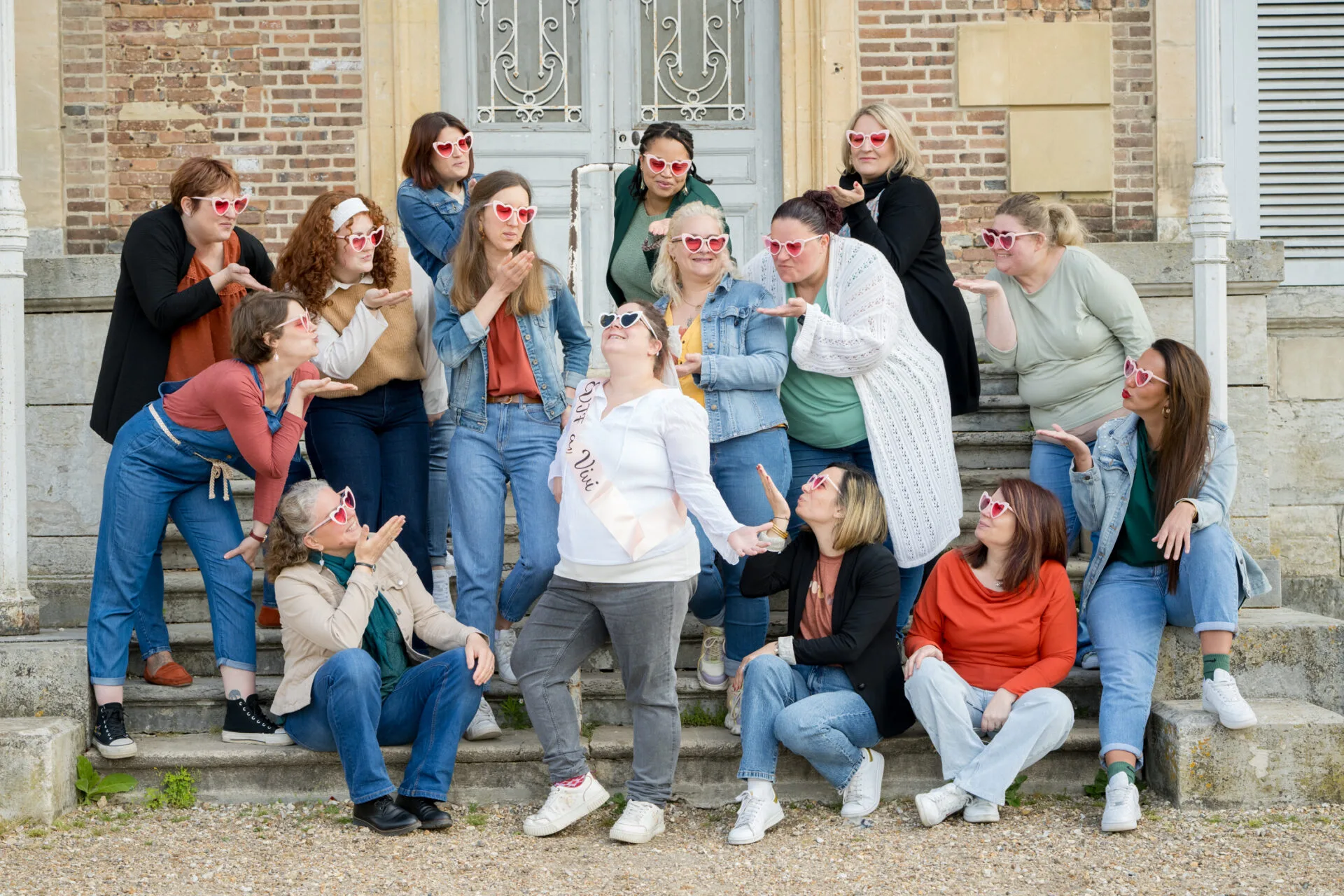 Groupe de femmes en lunettes cœur célébrant un EVJF avec joie, posant sur les marches d’un bâtiment à l’ambiance vintage