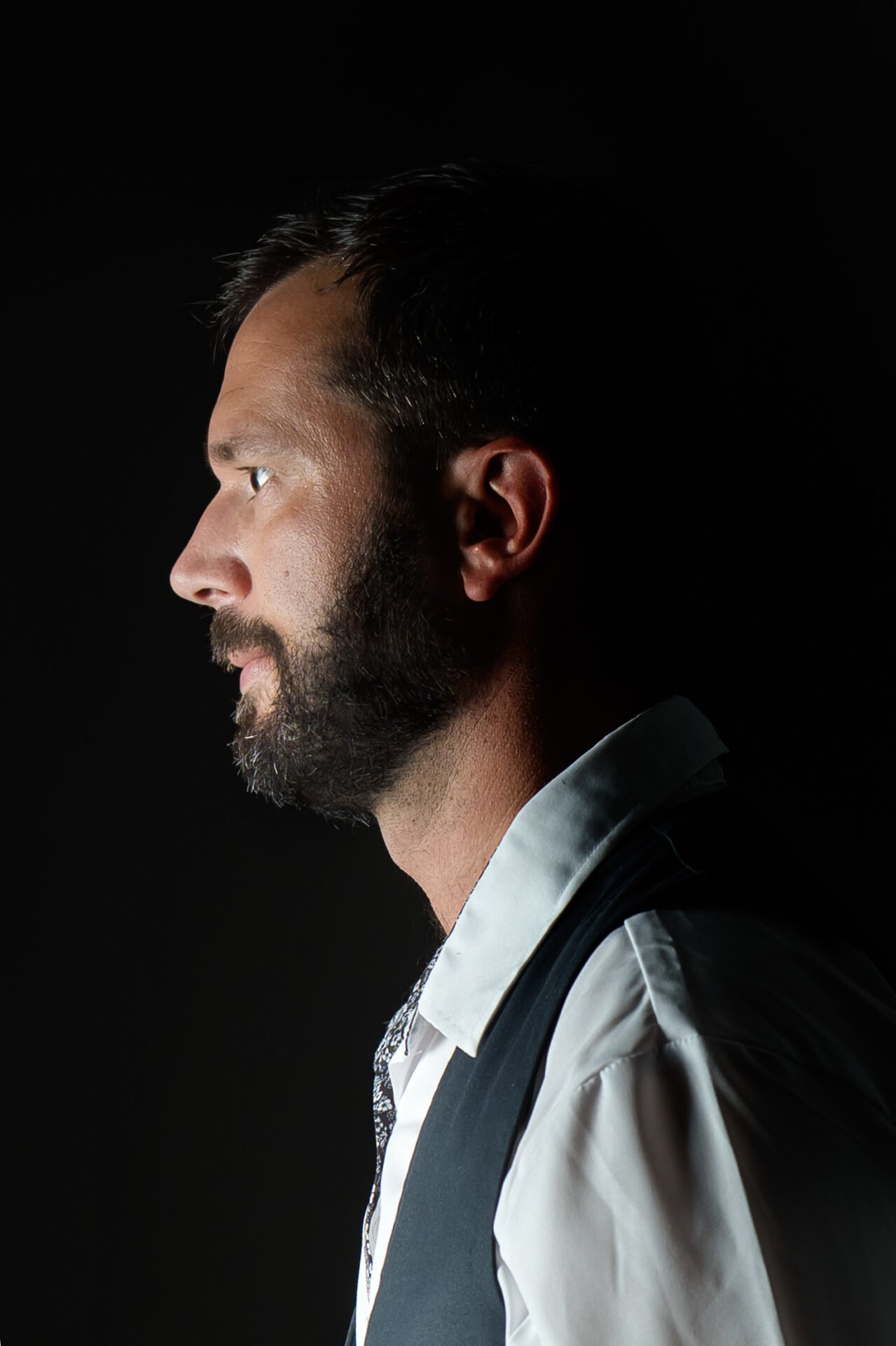 Homme barbu en chemise blanche et gilet noir posant les bras croisés, fond noir.