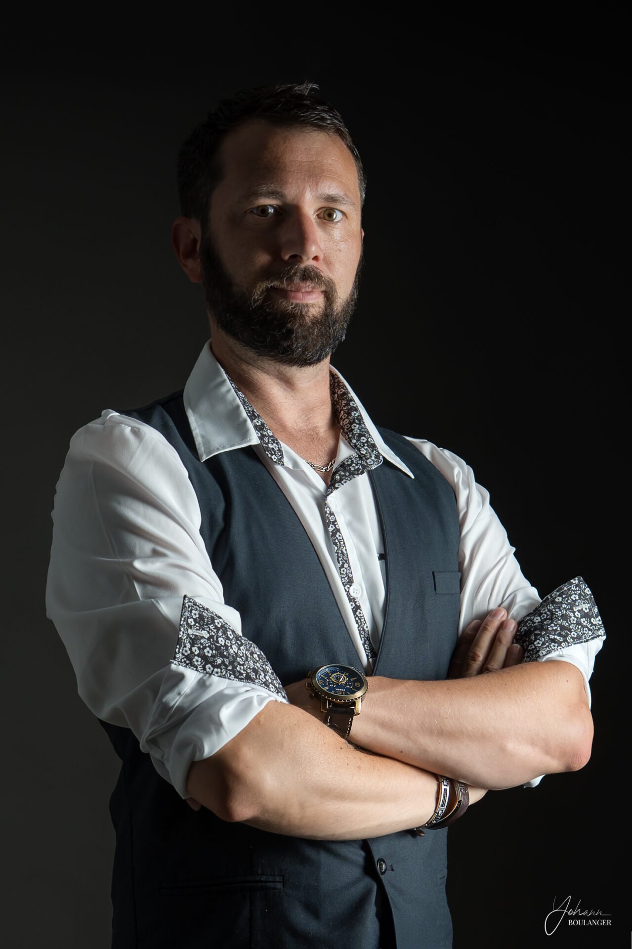 Homme barbu en chemise blanche et gilet noir posant les bras croisés, fond noir.
