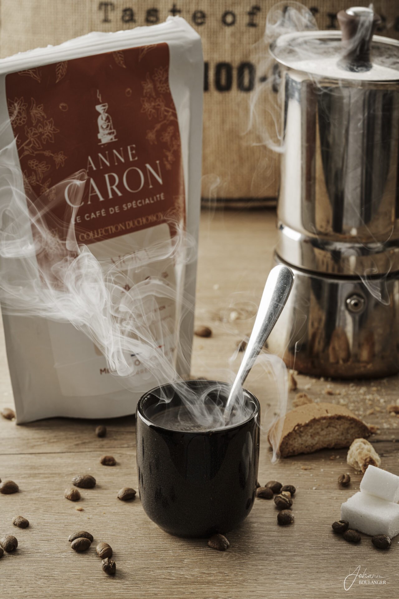 Photographie professionnelle d’un café haut de gamme Anne Caron avec tasse fumante, cafetière italienne et grains de café sur table en bois – prise de vue réalisée en Normandie